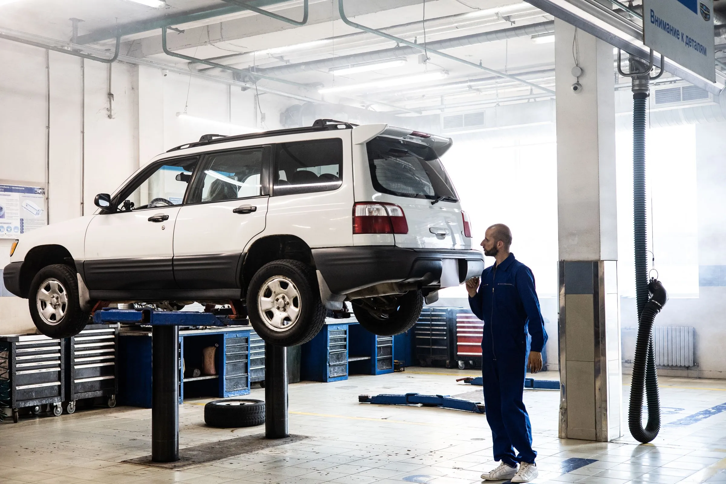 A technician in blue coveralls looking up at a white SUV raised on a lift in a bright, tidy workshop.