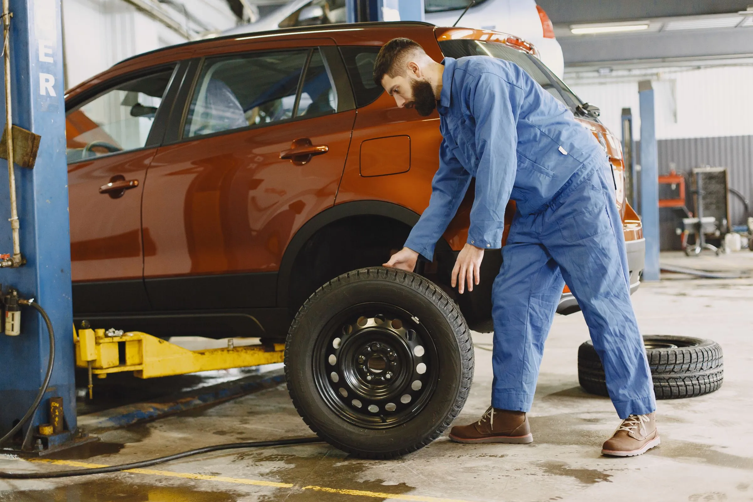 A mechanic in blue coveralls rolling a wheel and tire toward an orange SUV raised on a lift.
